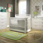 Grey baby nursery with white and beige patterned wallpaper, featuring a white crib, double dresser and 5 drawer dresser