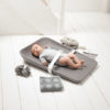 Baby strapped in with a white strap laying on a matty changing tray in grey surrounded by grey towels