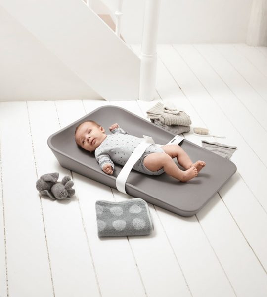 Baby strapped in with a white strap laying on a matty changing tray in grey surrounded by grey towels